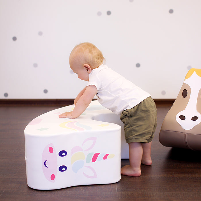 A baby standing next to a unicorn foam rocking toy